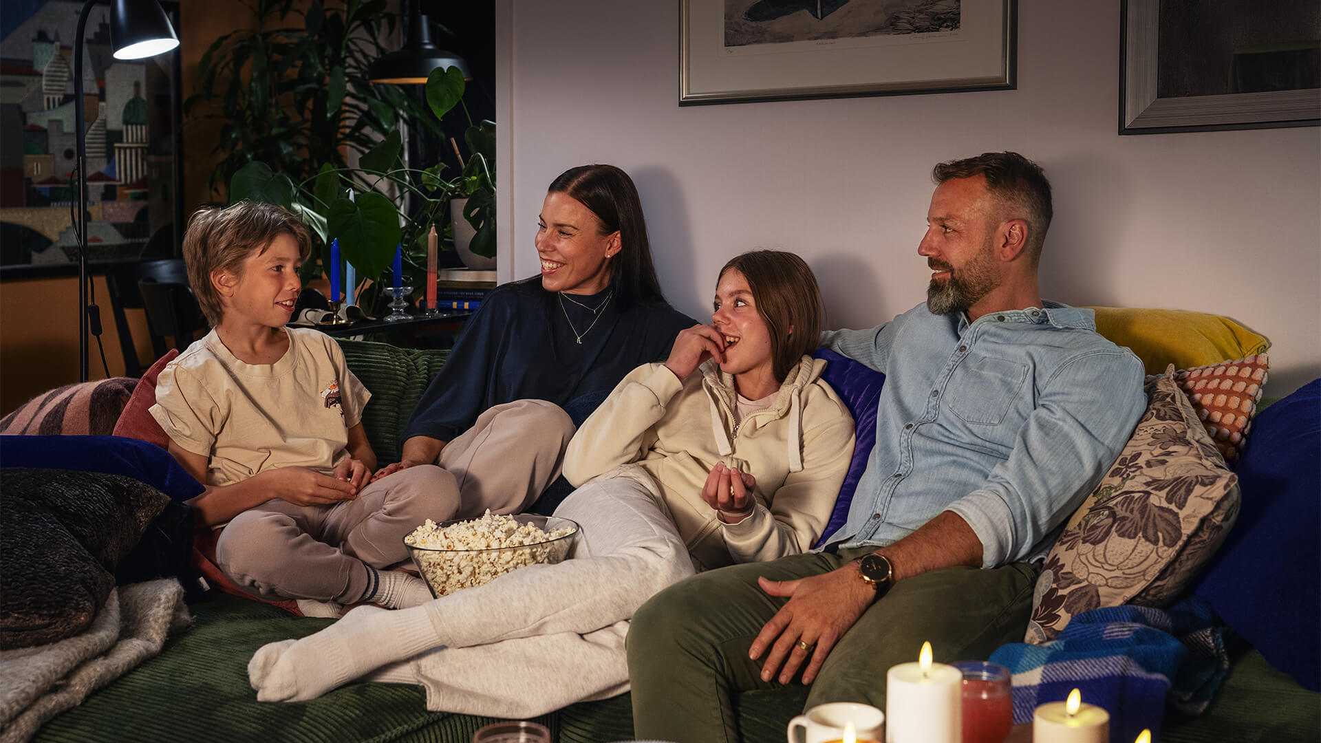 Neljä henkilöä — kaksi aikuista ja kaksi lasta — istuu sohvallla olohuoneessa hymyillen ja jutellen; keskellä on kulho popcorniä ja ympärillä tyynyjä, viltti ja kynttilöitä luomassa lämmintä, kodikasta tunnelmaa.